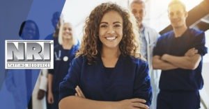 a close up of a mixed rave female nurse who is smiling and has her arm crossed, with other healthcare providers blurred in the background. They appear to be a team and everyone is smiling.