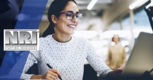 A female with black hair pulled back into a pony tail and wearing dark rimmed glasses is looking at a laptop screen and writing something down.