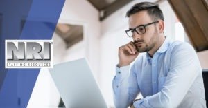 A white male wearing glasses and a while button down shirt is deep in thought and looking at a laptop screen