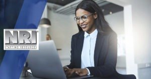 An African American women wearing grey round frame glasses is smiling whiel she types on a laptop computer. She is wearing a black blazer and white dress shirt.