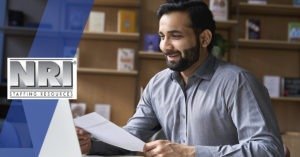 A man with black hair and a black beard is holding a piece of paper in his hands and smiling. He is wearing a grey button down shirt.