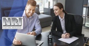 A Caucasian male is sittings down and pointing at a laptop computer, he is wearing a light blue button down shirt. a Caucasian female is leaning over to see what he is showing her. She is wearing a black blazer with a white button down shirt. She is holding a pen in her hands and is taking notes.