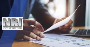 A close up of a pair of hands holding a piece of paper and a pen, this person also has a resume on the table and a laptop in the background