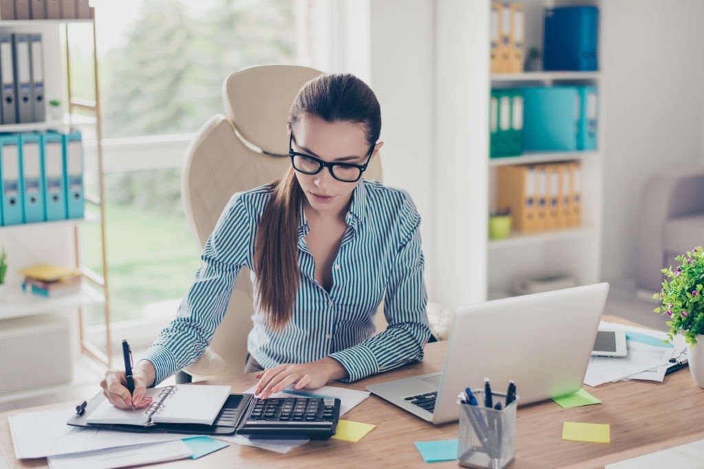 Portrait of serious young businesswoman accountant in formal wear and with pony tale, sitting at her work place and doing notes
