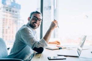 Portrait of cheerful male entrepreneur in classic eyewear smiling at camera while working at desktop table with modern laptop computer,